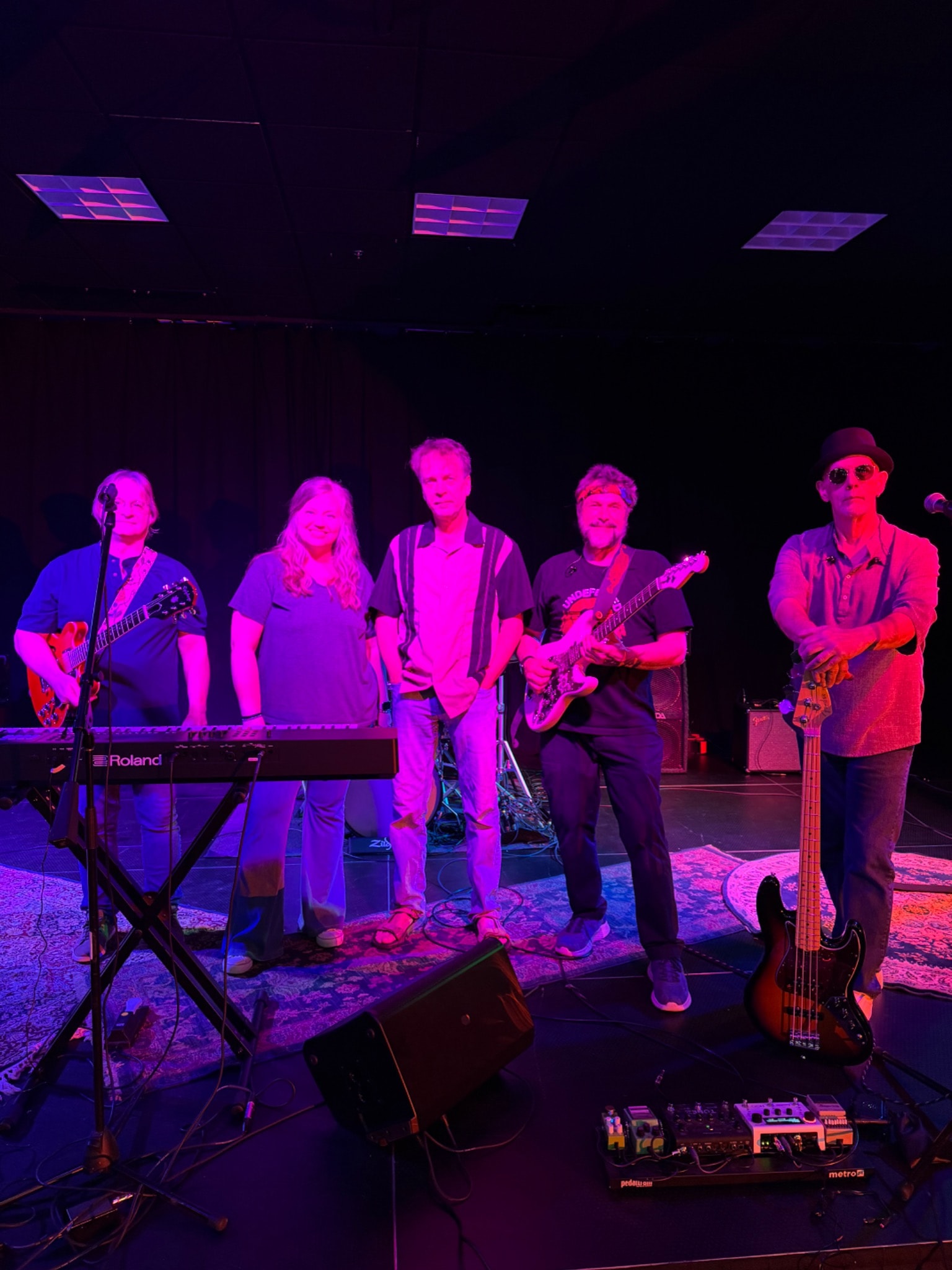 Five musicians standing together with guitars, bass, and a keyboard on stage.